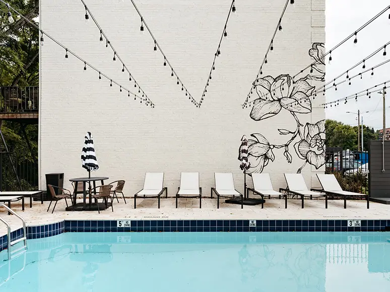 Pool deck with lounge chairs, umbrellas, and string lights beside a white brick mural wall