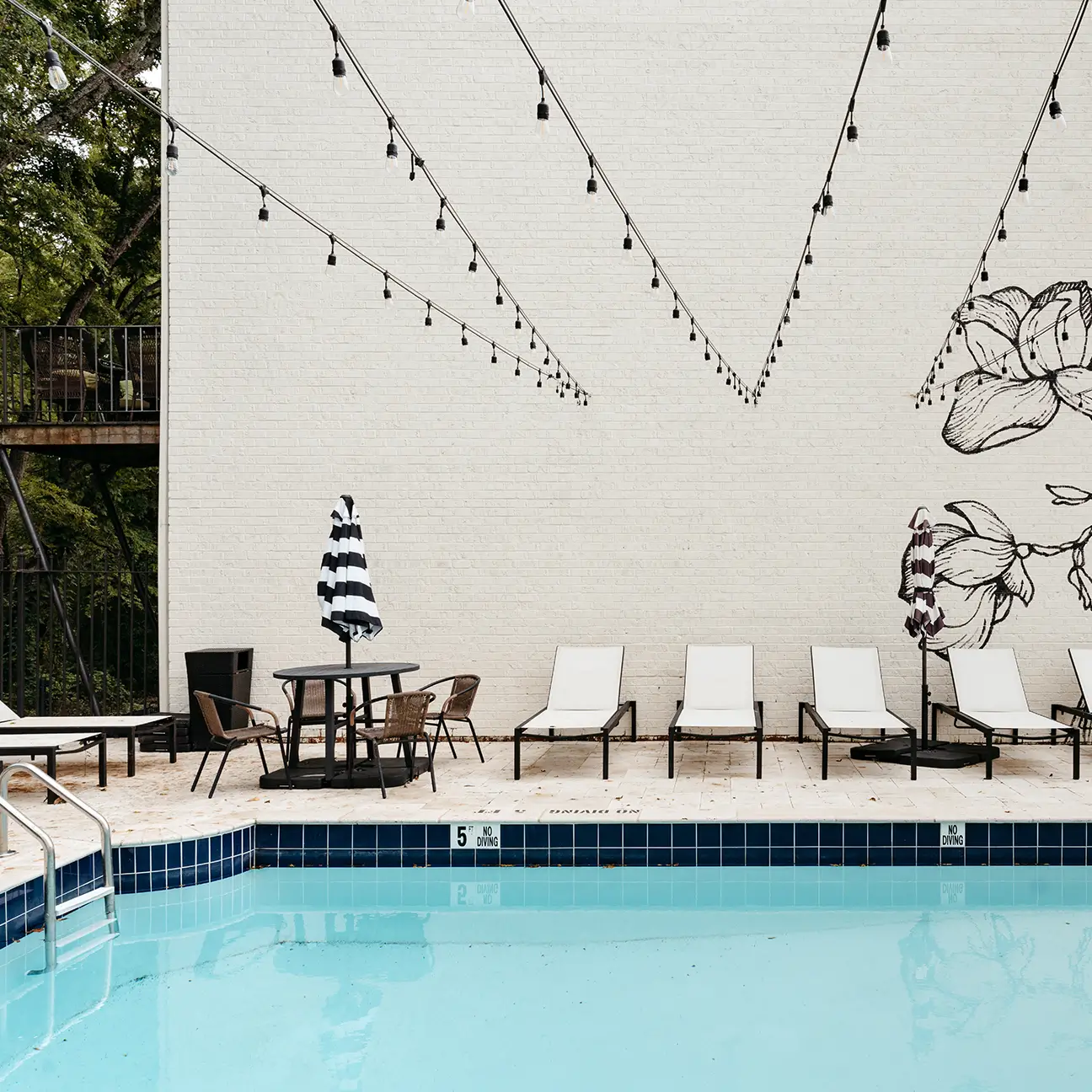 Pool deck with lounge chairs, umbrellas, and string lights beside a white brick mural wall